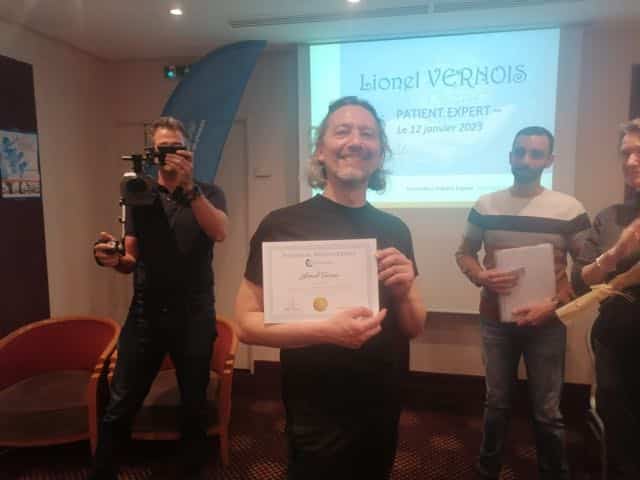 Homme souriant tenant un certificat lors d’une remise de prix en salle, projection affichant « Lionel VERNOIS » à l’arrière.